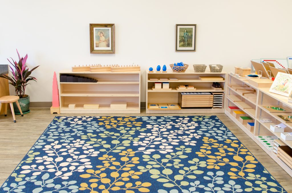 A bright Montessori Children’s House classroom with low wooden shelves displaying Sensorial materials such as the Pink Tower, Brown Stair, knobbed cylinders, geometric solids, and color tablets. A large patterned rug covers the floor, and framed artwork hangs on the wall above the materials.