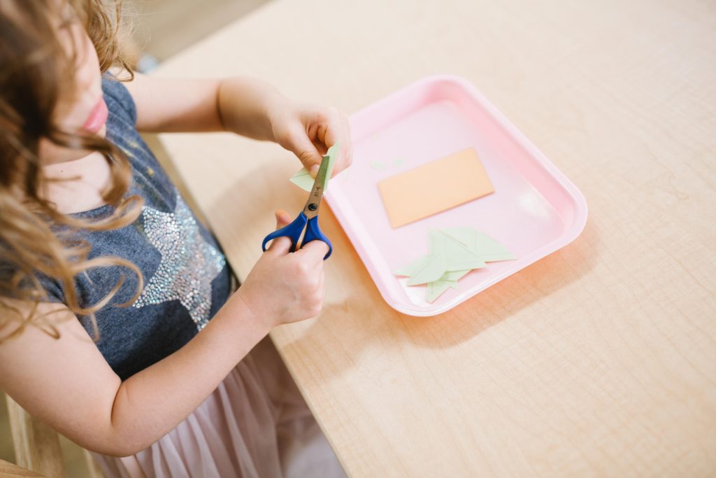 A young Montessori student uses scissors to cut green paper shapes on a pink tray, practicing early scissor skills.