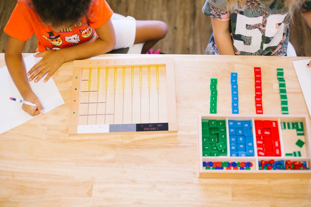 Overhead view of children working with the Montessori Stamp Game, arranging green, blue, and red place value tiles and writing answers on graph paper.