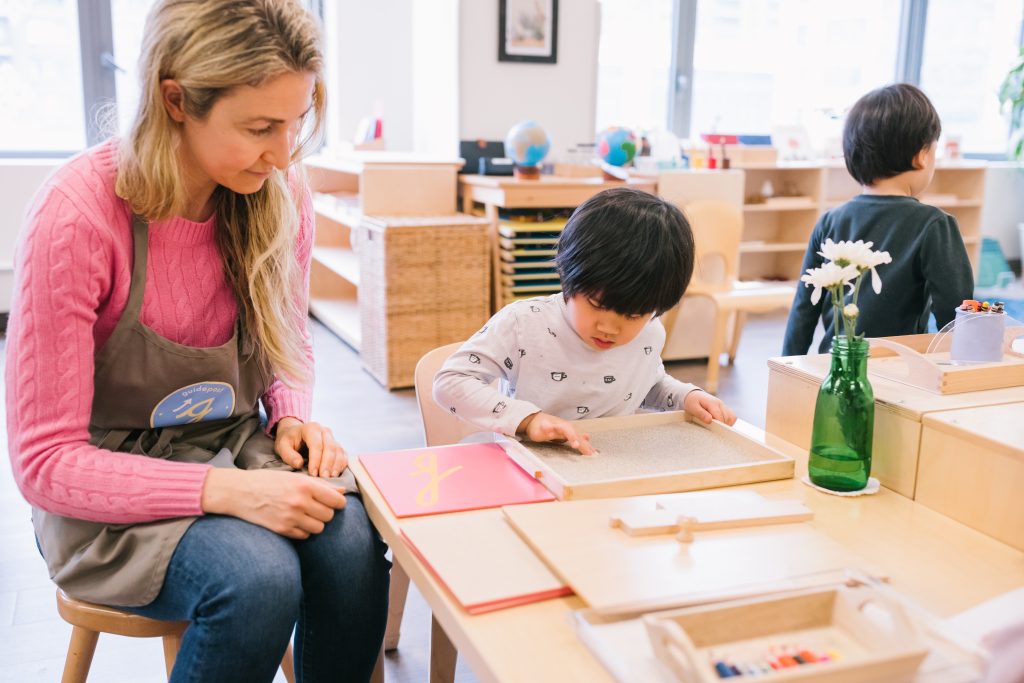 Guidepost Montessori guide sitting beside a child working with a sandpaper letter at a classroom table.