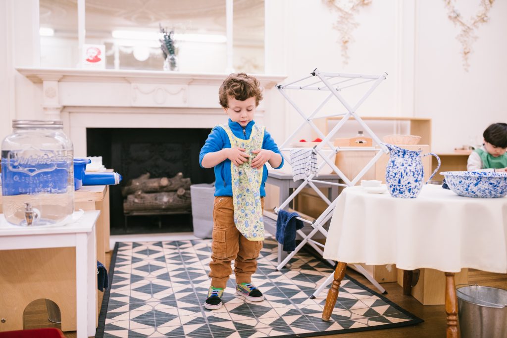 Young child in a Montessori classroom carefully folding a cloth as part of a practical life activity. Drying rack and water materials are visible in the background.