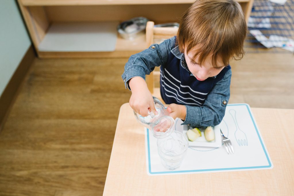 Young child concentrating while pouring water from a small glass pitcher during a Montessori snack routine.