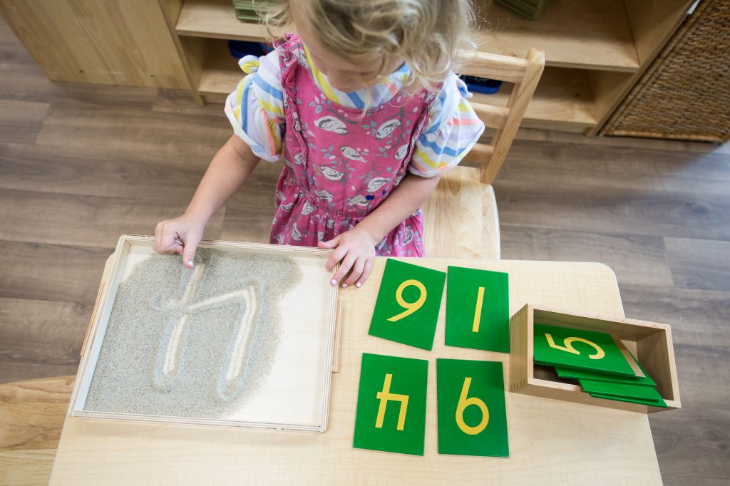 Child tracing a number in a Montessori sand tray beside green number cards on a table.