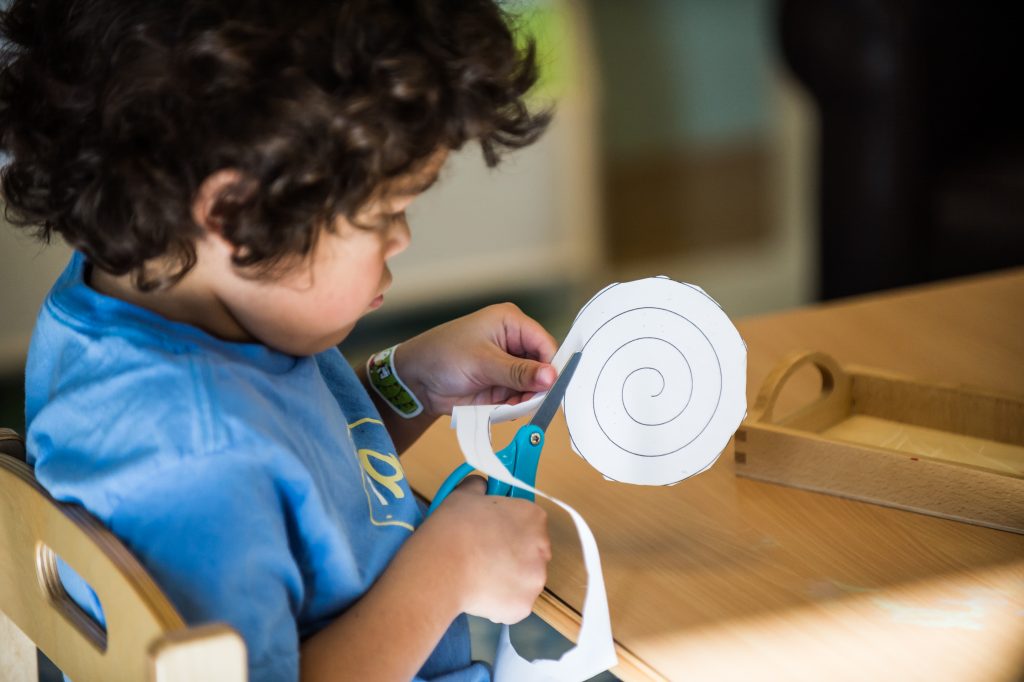 A Montessori child cuts along a spiral paper pattern using blue scissors during a fine motor activity.