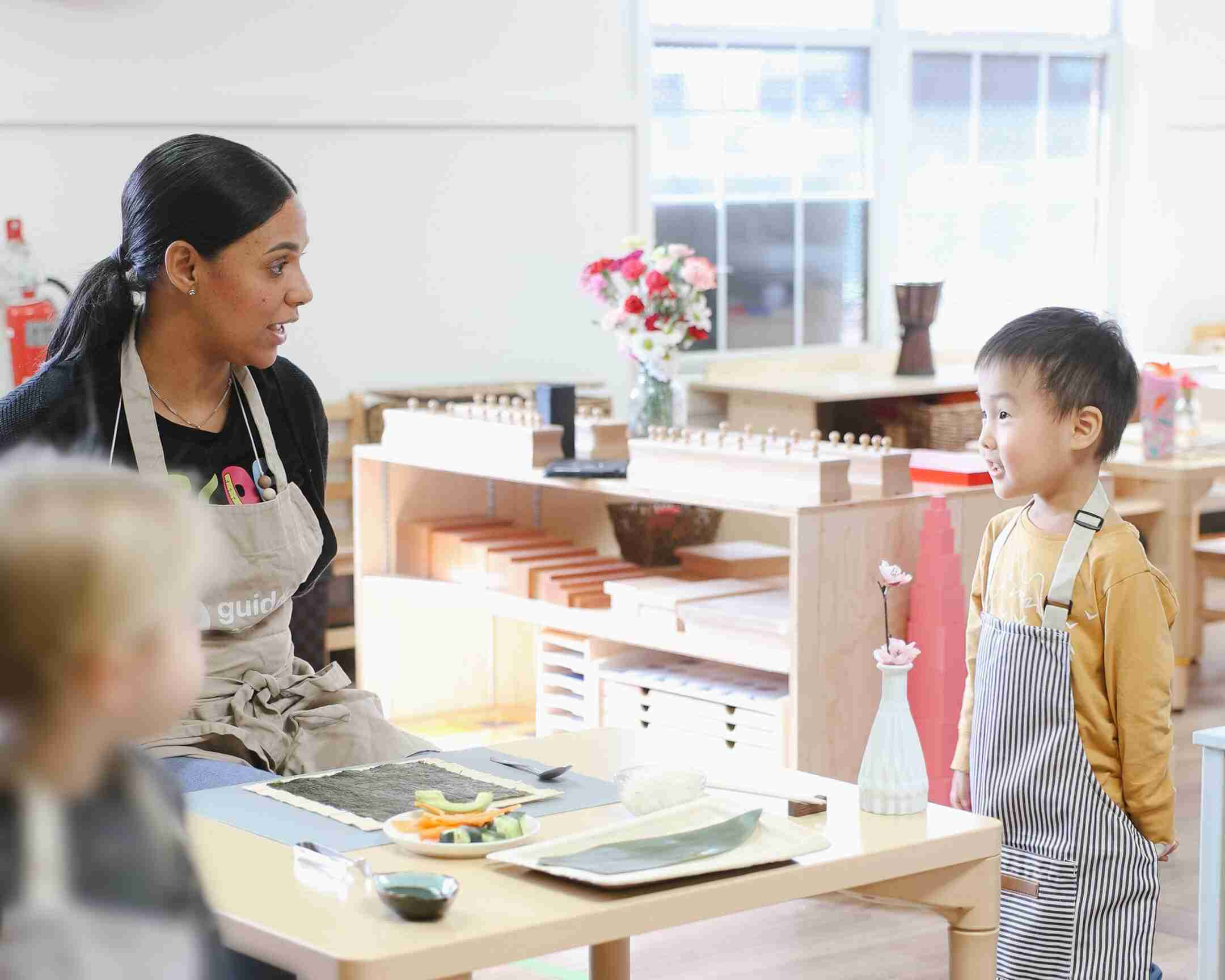 Guide at Guidepost Montessori Mahwah speaks with a smiling preschooler in an apron beside a sushi-making setup during a cultural practical life lesson.