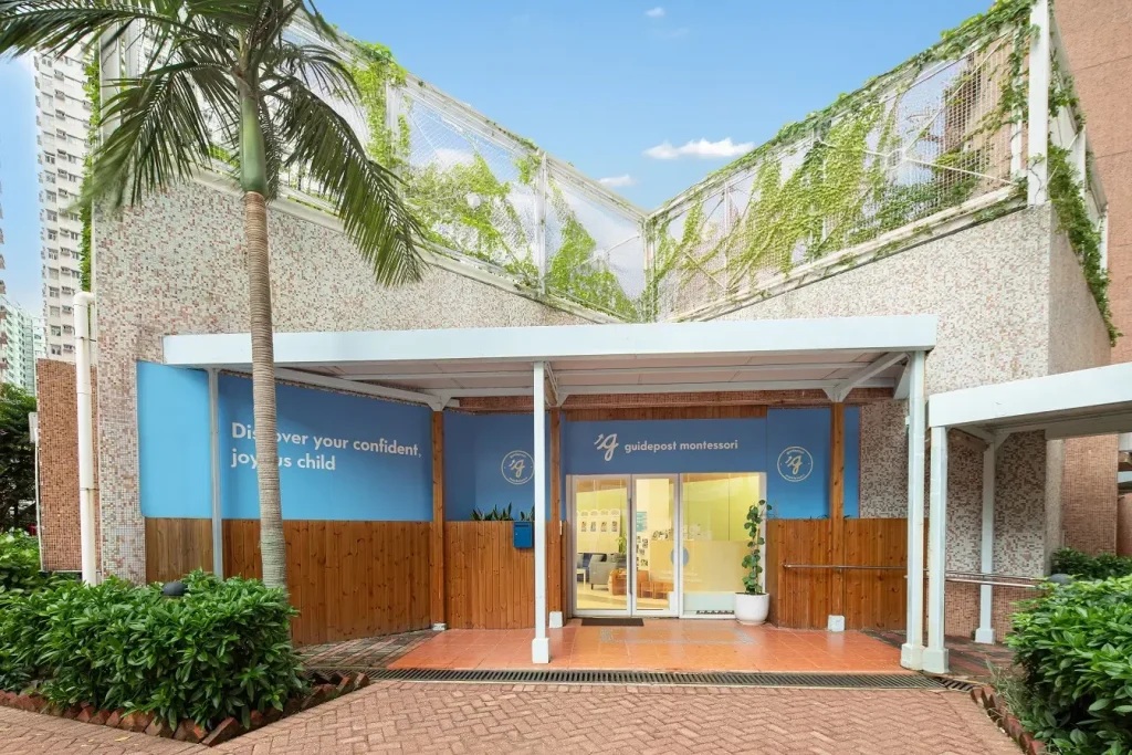 Exterior view of a Guidepost Montessori school entrance with blue branding, wooden fencing, and greenery, creating a welcoming environment for families.