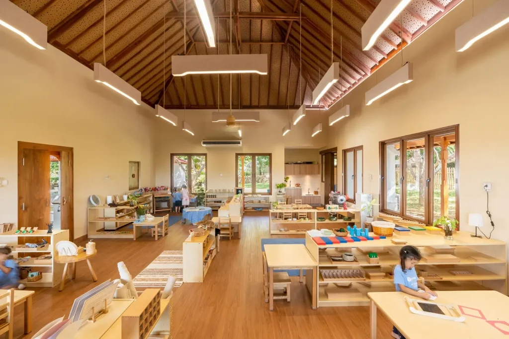 Spacious Montessori classroom in Bali with wooden furniture, natural light from large windows, and children engaged in hands-on learning activities.